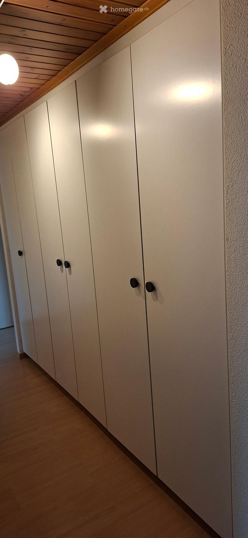 A narrow hallway with white built-in cabinets and a wooden ceiling. The floor is light wood, and the walls are textured white.