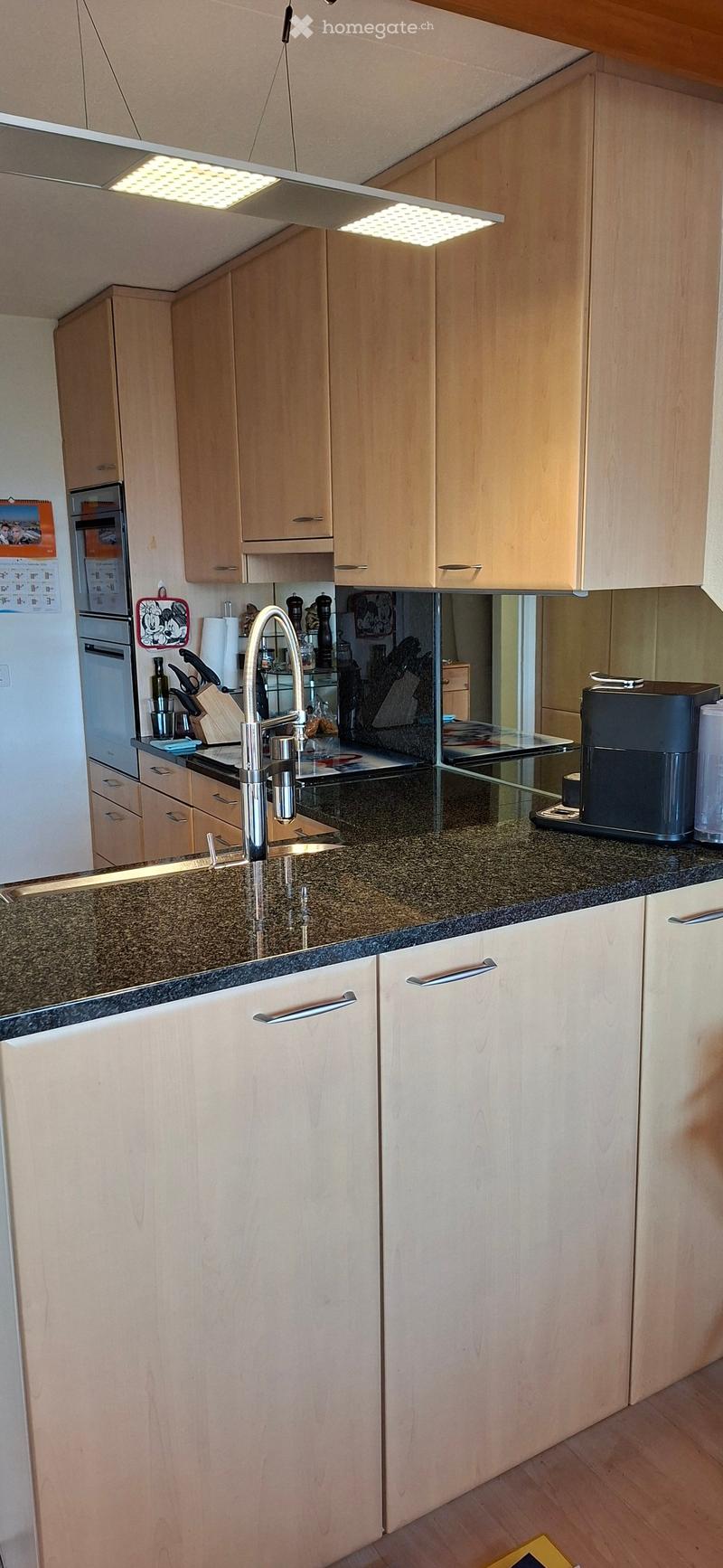 Modern kitchen with light wooden cabinets, granite countertop, stainless steel faucet, coffee maker, and various kitchen appliances.