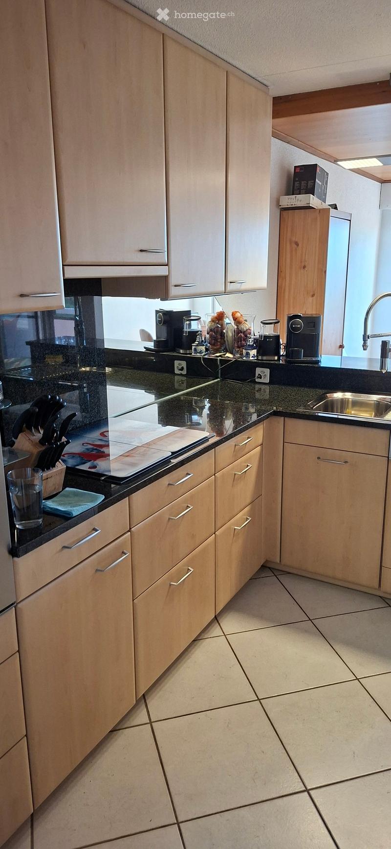 Modern kitchen with light wood cabinets, granite countertops, and stainless steel sink. Coffee maker, blender, and other appliances are on the counter.