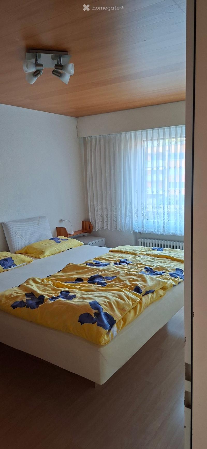 Cozy bedroom with a double bed covered in a yellow and blue floral duvet, a wooden nightstand with a lamp, and white curtains on the window.