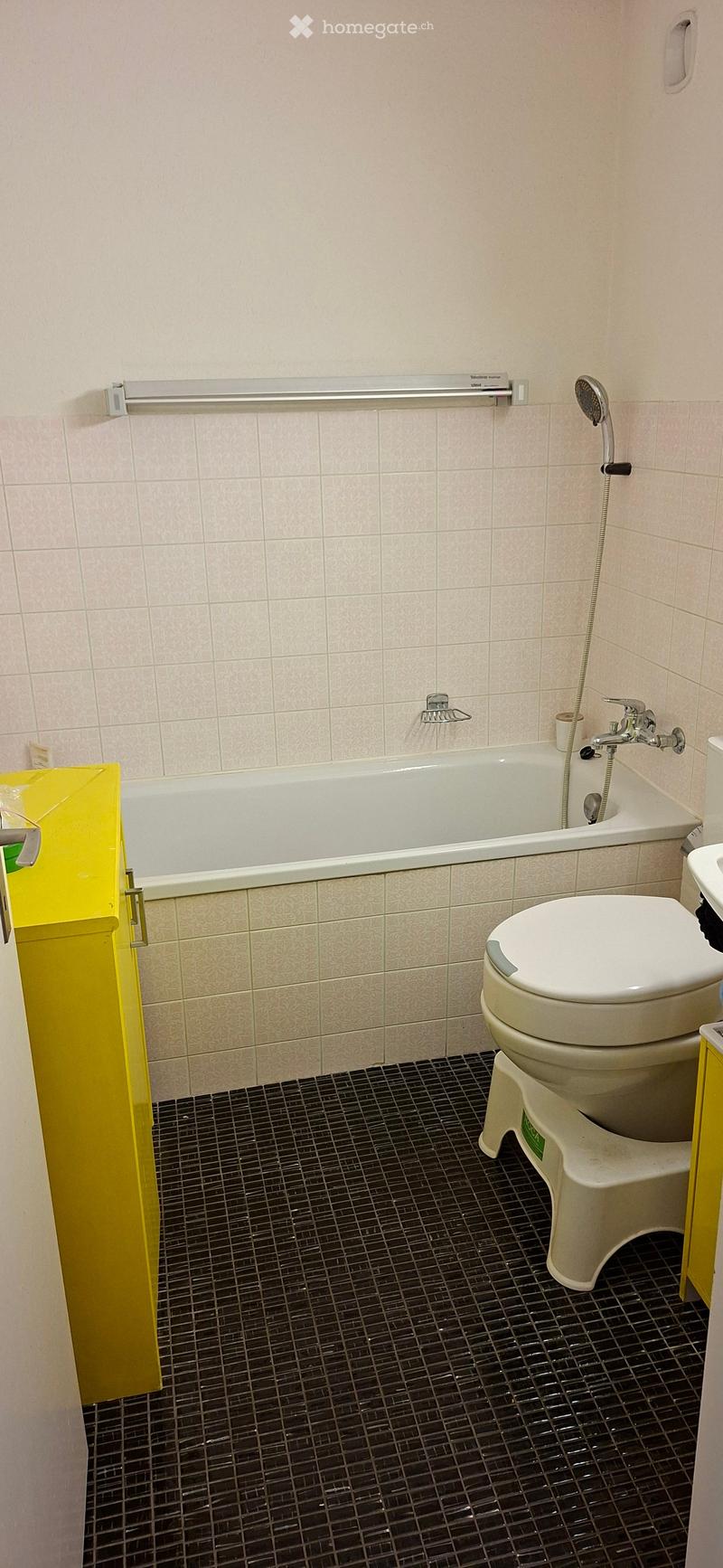 Bathroom with a bathtub, toilet, showerhead, and faucet. The walls are tiled with a light pink pattern, and the floor has dark black tiles.
