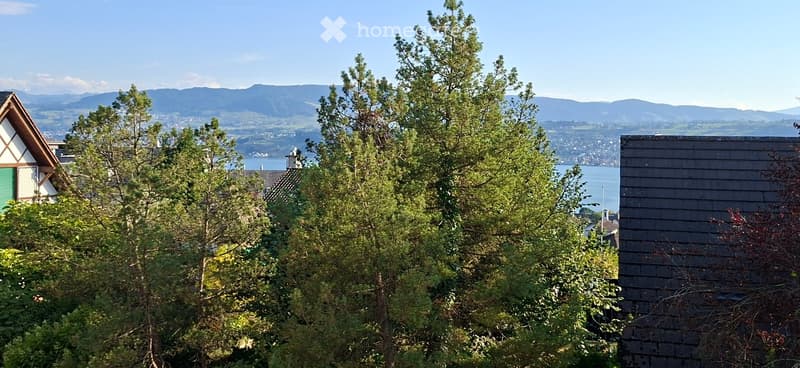 View of a house and surrounding landscape with trees, mountains, and a body of water in the background.