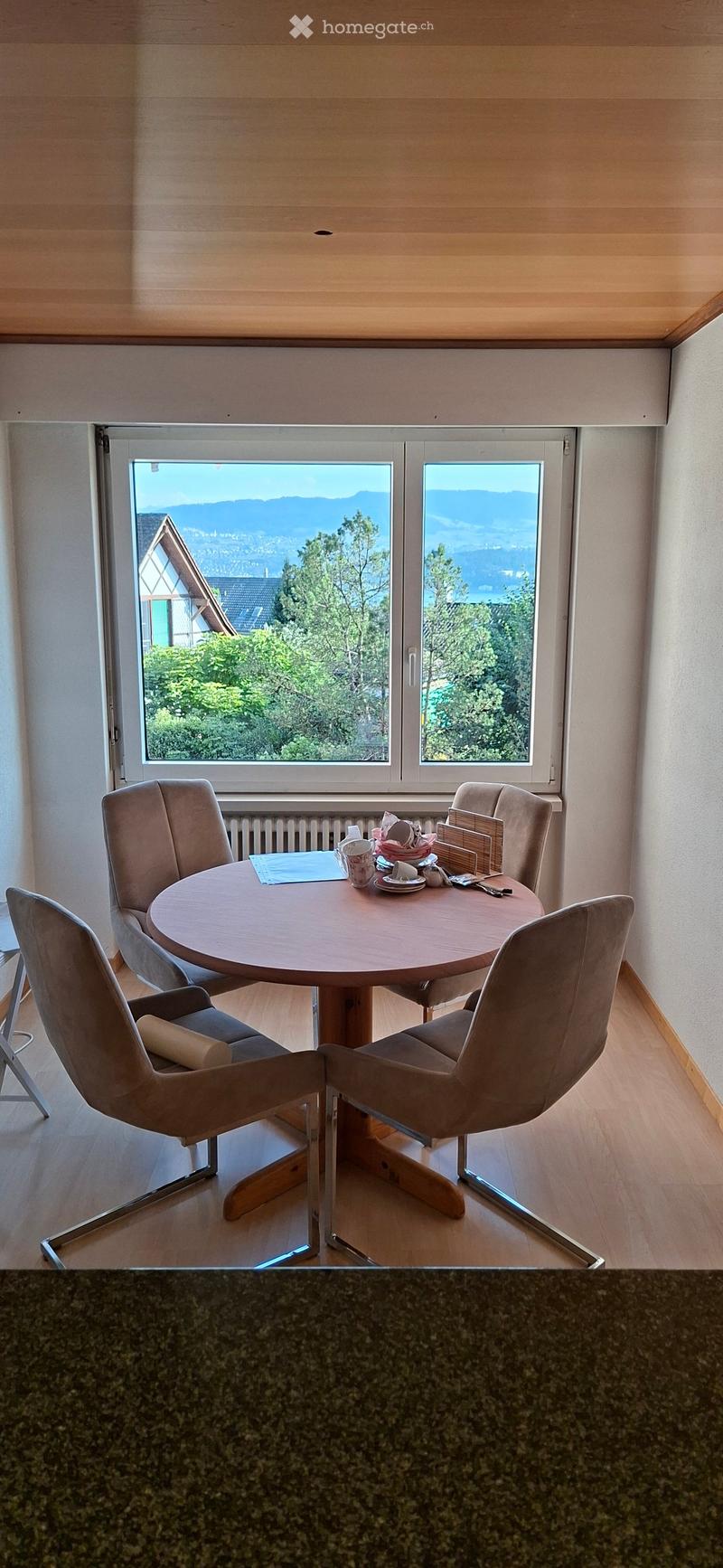Dining area with a round wooden table, four beige chairs, and a scenic view of mountains and greenery through large windows.