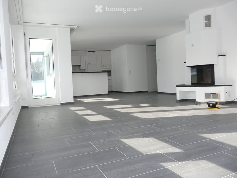 Empty room with grey tiled floor, white walls, and large windows allowing natural light. Features a fireplace and a kitchen area with cabinets and a countertop.