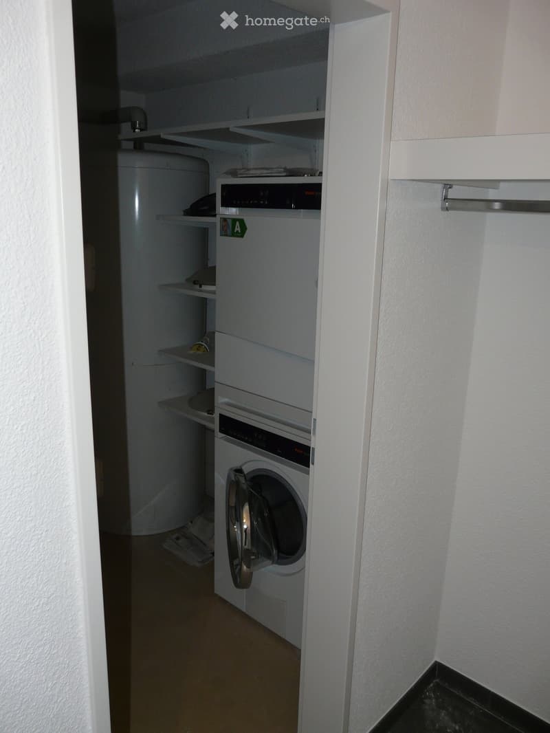 A small utility area with a washing machine, dishwasher, and refrigerator, featuring white walls and shelves.
