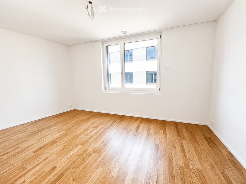 Empty room with light wooden flooring and white walls, featuring a large window with white frame and blinds.