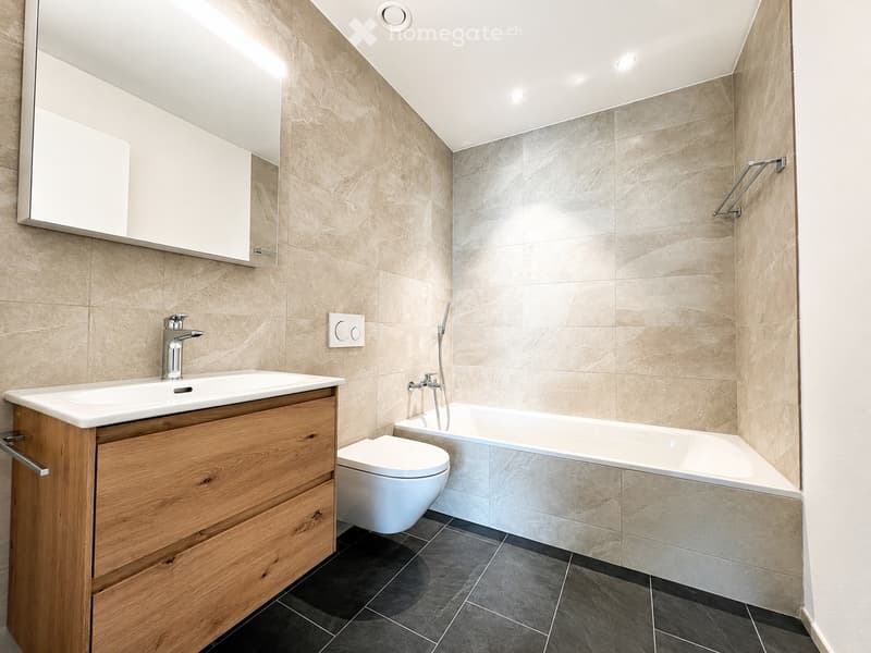 Modern bathroom with beige tiled walls, a wooden vanity with a sink, a bathtub, and a toilet. Features a large mirror above the sink and a shower rail in the corner.