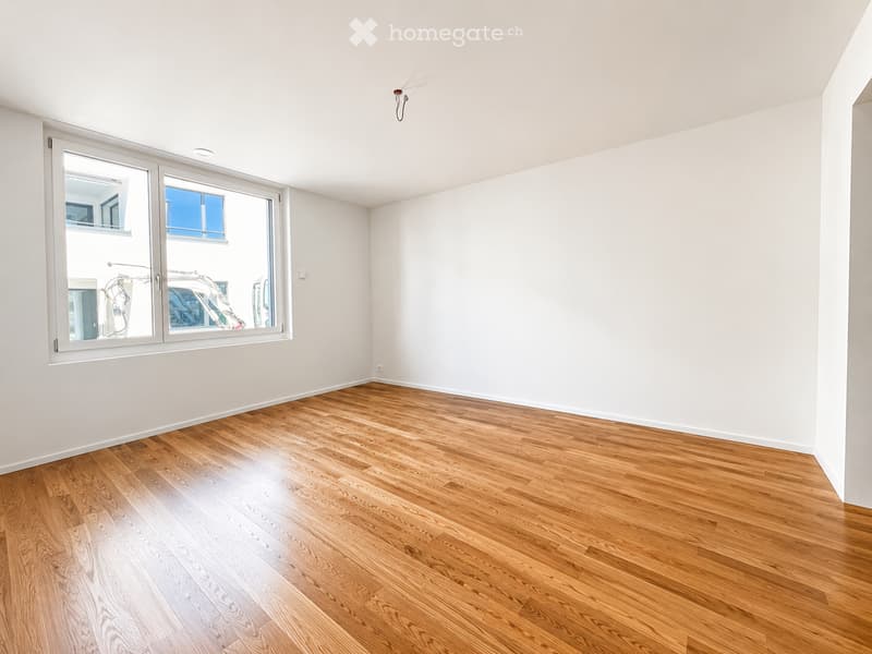 Empty room with light-colored wooden flooring and white walls, featuring large windows with a view of the outside.