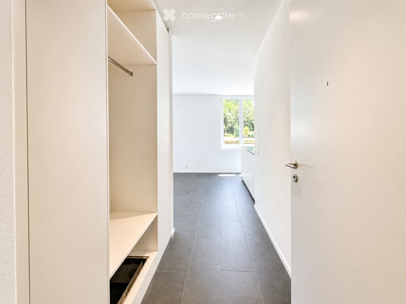 Modern hallway with grey tiled floor, white walls, built-in storage unit, and a large window at the end showing greenery outside.