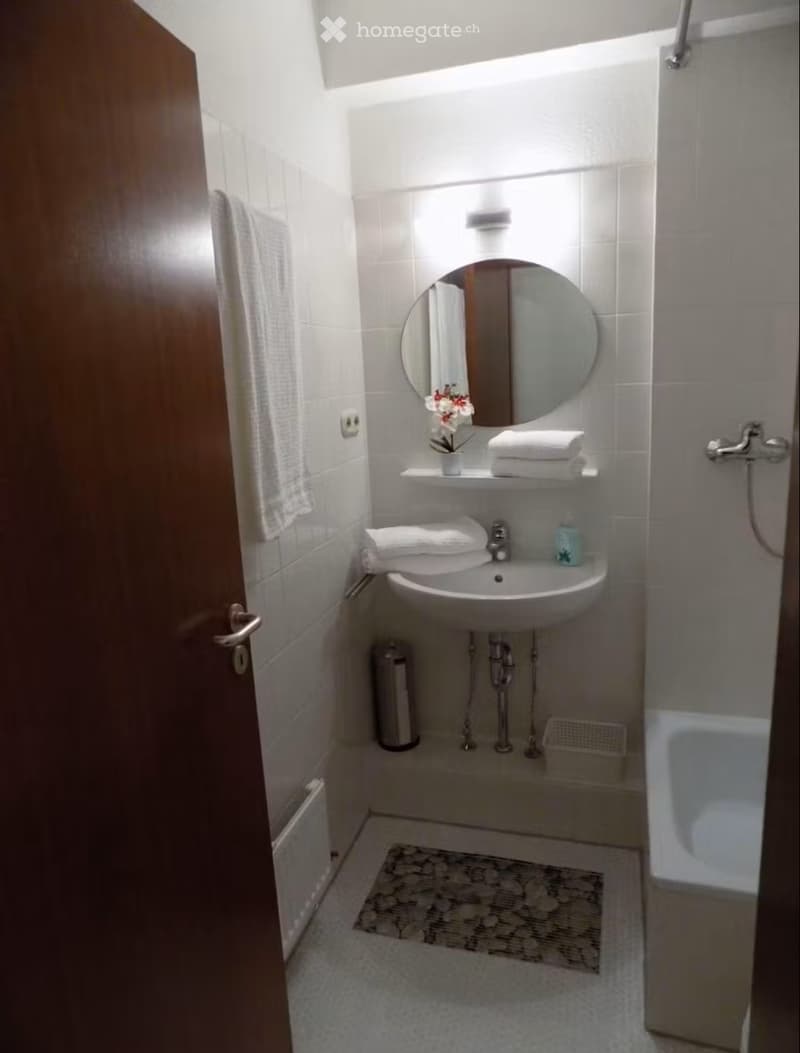 Modern bathroom with white tiles, a round mirror, a sink with a faucet, neatly folded towels, a small flower vase, and a bathtub.