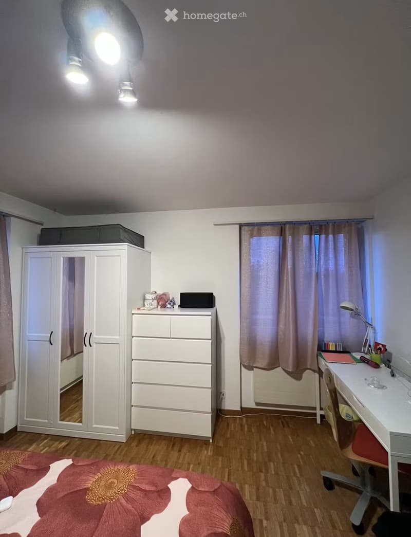 Well-lit bedroom with wooden flooring, a large wardrobe, a drawer unit, a desk with a chair, and a bed with a floral-patterned bedspread.