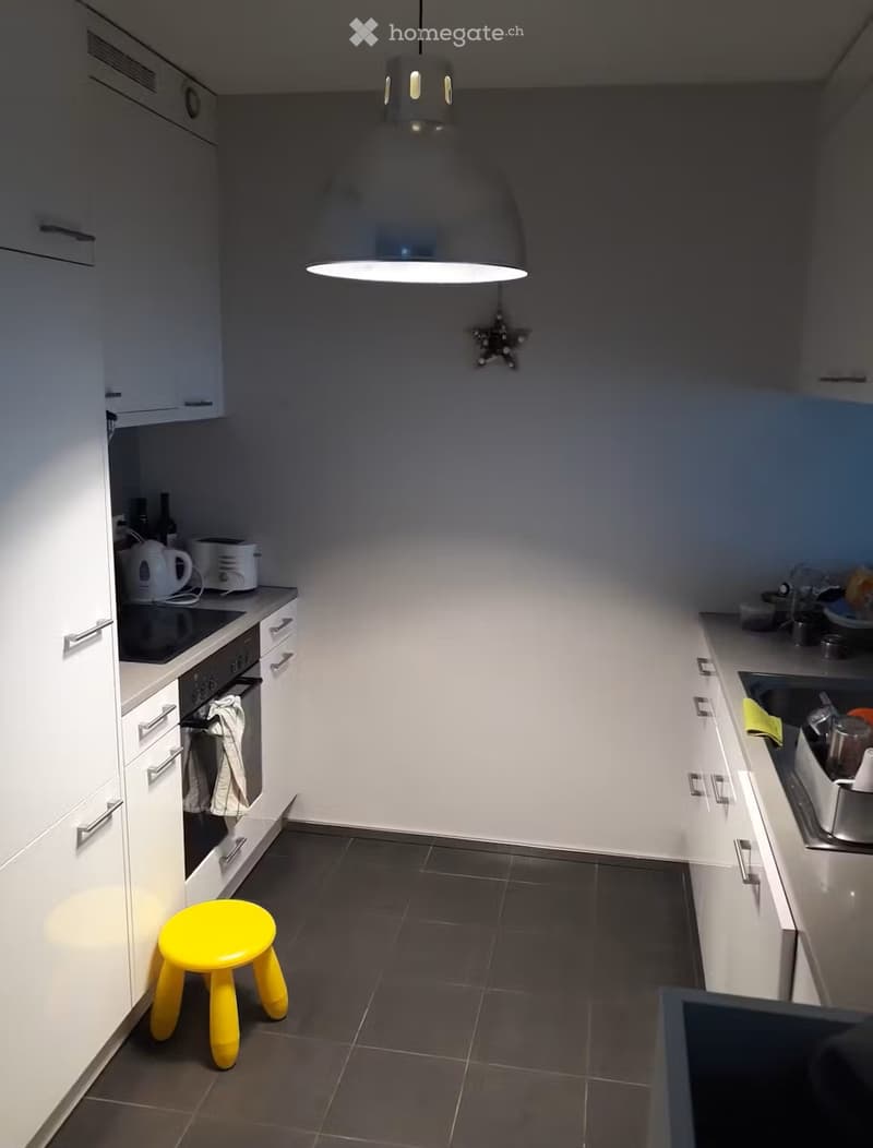 Modern kitchen with white cabinets, dark tiled floor, and a yellow stool in the center. Features an oven, sink, and various kitchen appliances on the counters.