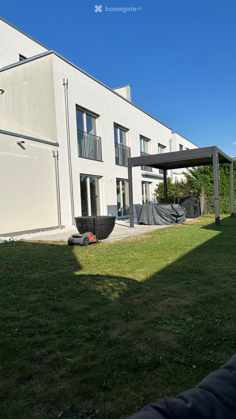 Well-maintained lawn in front of a modern white house with large windows and balconies. A covered patio area with outdoor furniture is visible to the right.