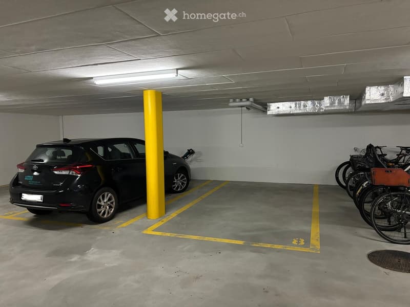 Underground parking area with a parked car and several bicycles, marked parking spaces, and a yellow support pillar.