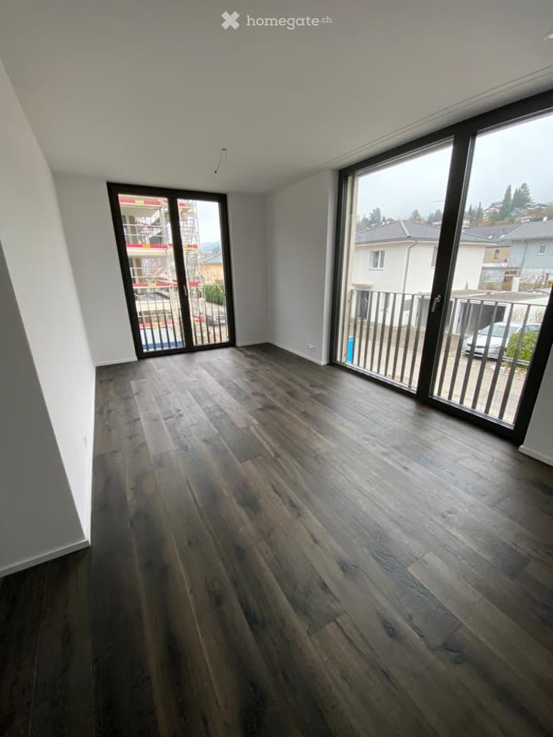 Empty room with dark wooden flooring, large sliding glass doors leading to a balcony, white walls, and modern lighting fixtures.
