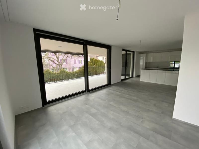 Empty room with large sliding glass doors leading to an outdoor area, grey tiled floor, white walls, and a view of a garden and neighboring house.