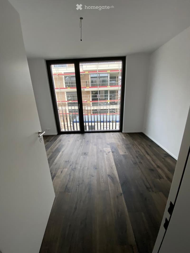 Empty room with wooden flooring, large glass sliding door leading to a balcony, white walls, and a visible ceiling light fixture.