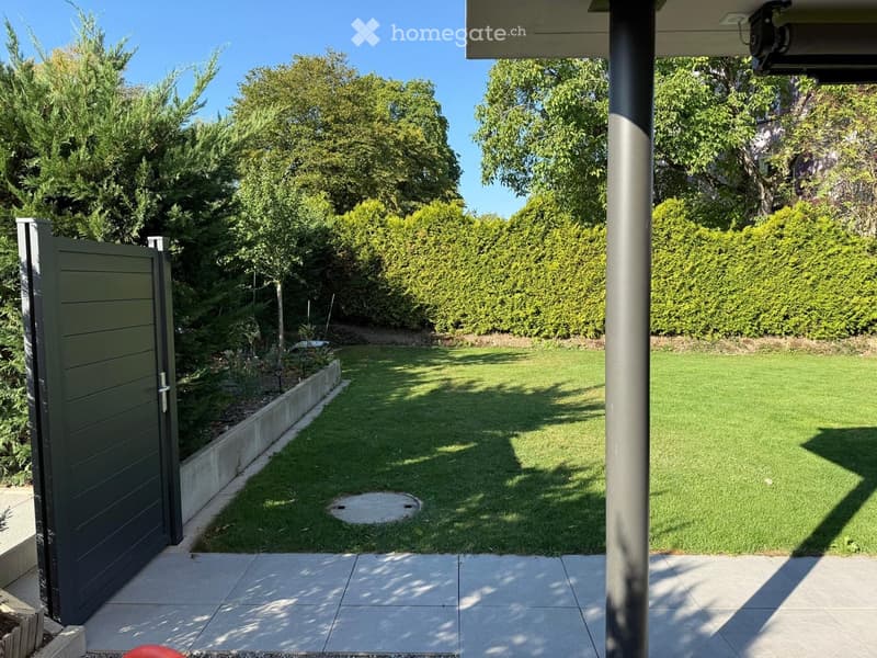 Well-maintained garden with a neatly trimmed lawn, a concrete pathway, and surrounding greenery including trees and shrubs.