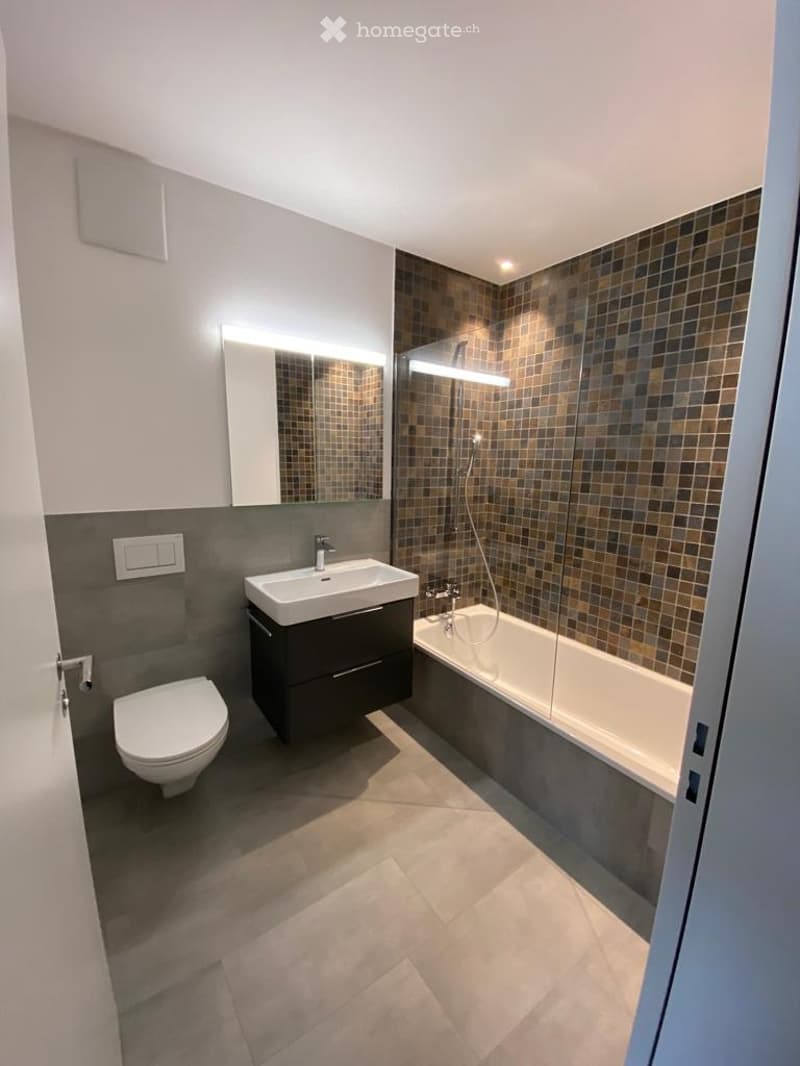 Modern bathroom with a white toilet, a dark wooden sink cabinet, a bathtub with a glass shower enclosure, and a large mirror with built-in lighting.