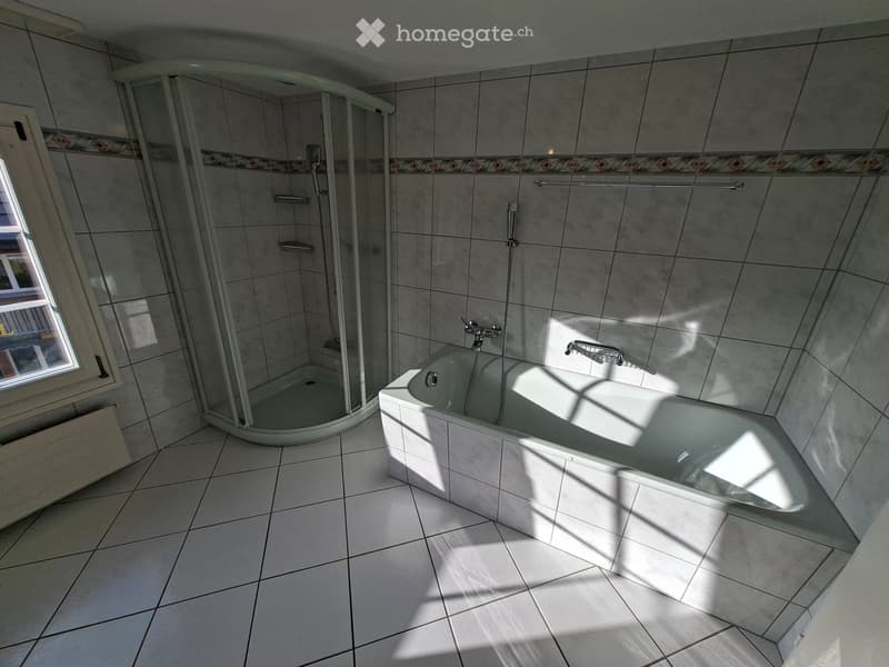 Modern bathroom with a bathtub, glass-enclosed shower, and tiled walls and floor. Sunlight streams in from a window on the left.