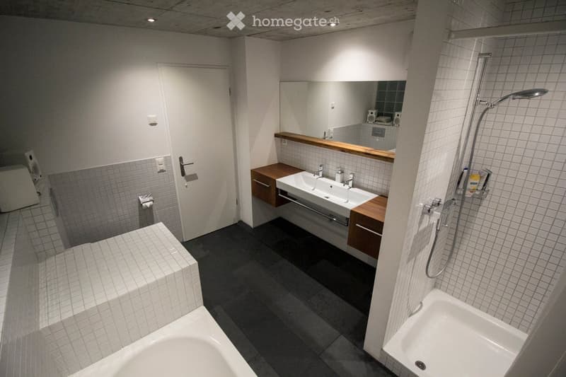 Modern bathroom with a double sink, bathtub, shower, and built-in wooden cabinetry. The space features tiled walls and a dark floor, with a large mirror above the sink.