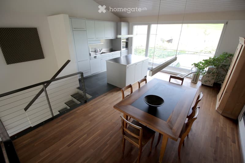Open-plan kitchen and dining area with light-colored cabinets, a central island, and a wooden dining table with chairs. The space features wooden flooring and large windows with a view of a balcony.