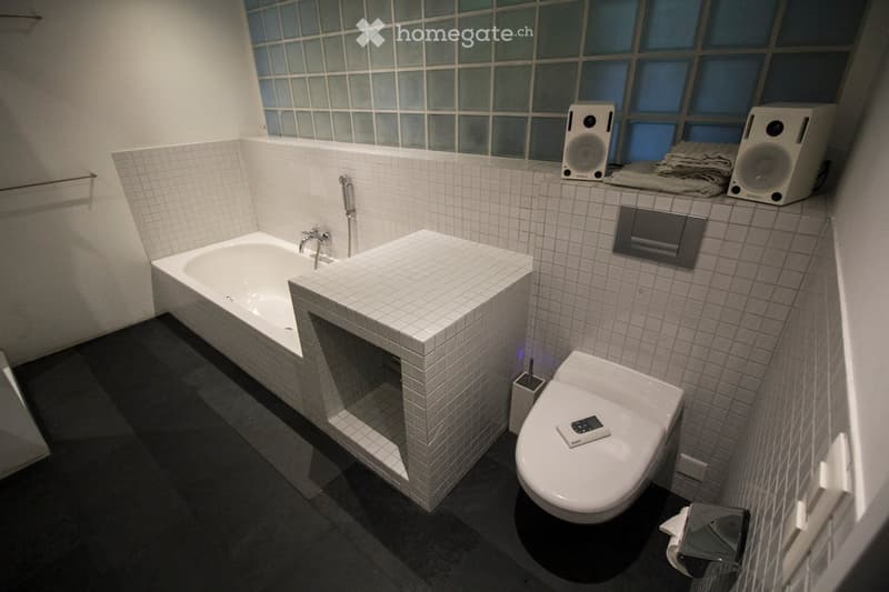 Modern bathroom with a bathtub, toilet, sink, and speakers on the counter. The walls are tiled, and there is a remote control on the toilet seat.