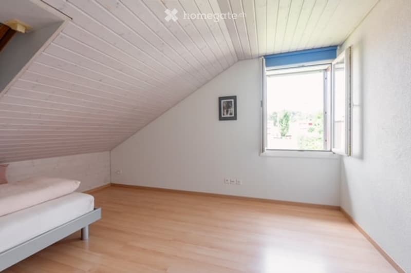 Bright, minimalist bedroom with wooden flooring, white walls, slanted ceiling, and a single bed against one wall. A framed picture hangs on the wall, and a window with white frames lets in natural light.