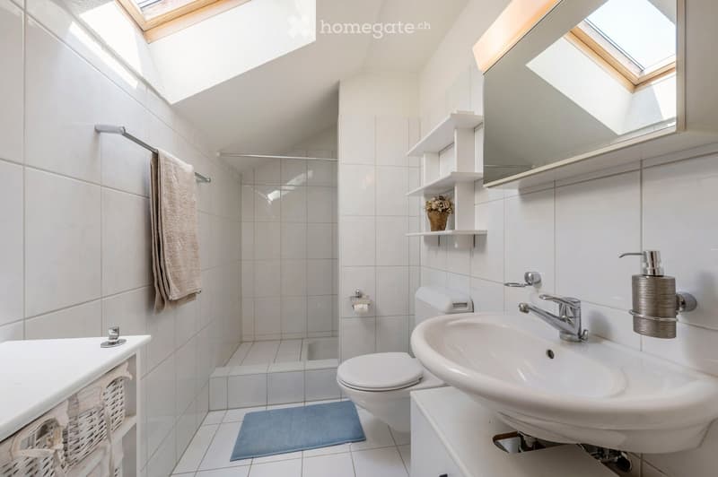 Modern bathroom with white tiles, a sink, toilet, mirror, towel, and a small shelf with a flower vase.