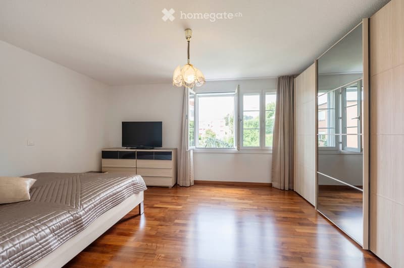 Modern bedroom with wooden flooring, a large bed with a dark duvet, a TV on a wooden stand, and a large mirrored wardrobe.