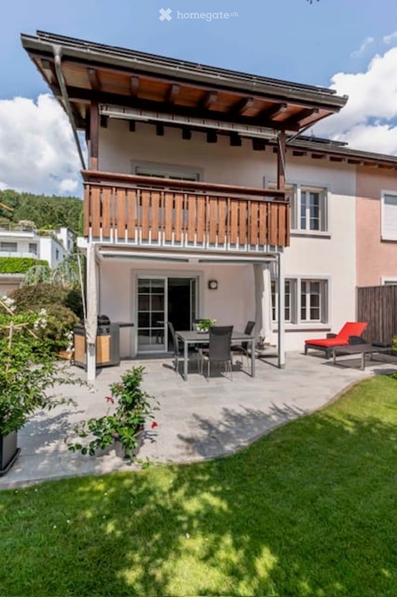 A modern house with a wooden balcony, patio area with a dining set, lounge chairs, and a well-maintained lawn.
