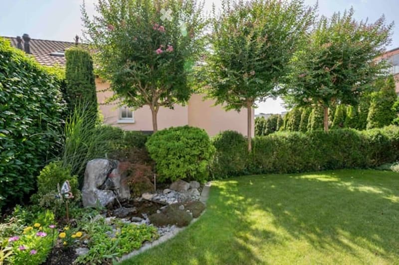 Well-maintained garden with a small water feature, various shrubs, trees, and a neatly trimmed lawn.