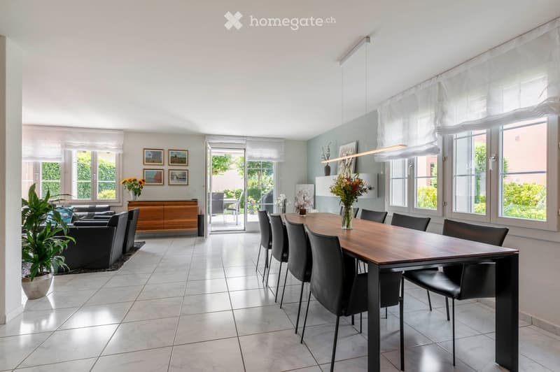 Bright dining area with a long wooden dining table surrounded by black chairs, a black leather sofa, a wooden cabinet, and decorative vases. Large windows and sliding glass doors lead to a patio.