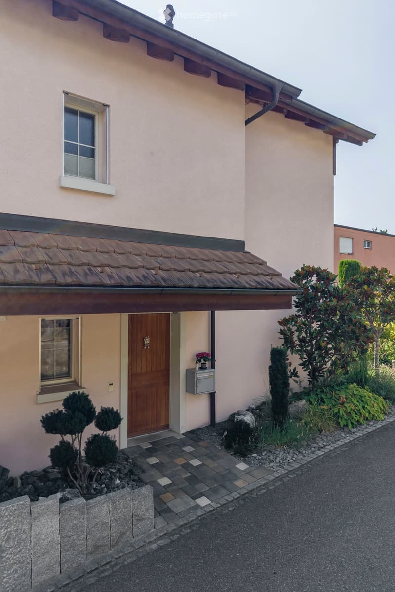 Single-story house with light pink exterior, wooden front door, small porch, and neatly trimmed shrubs.