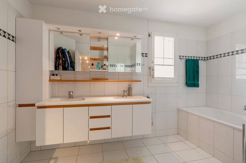 Modern bathroom with white tiles, double sink, large mirror, and a bathtub. Features include a window, a towel hanging on the wall, and a cabinet with wooden accents.