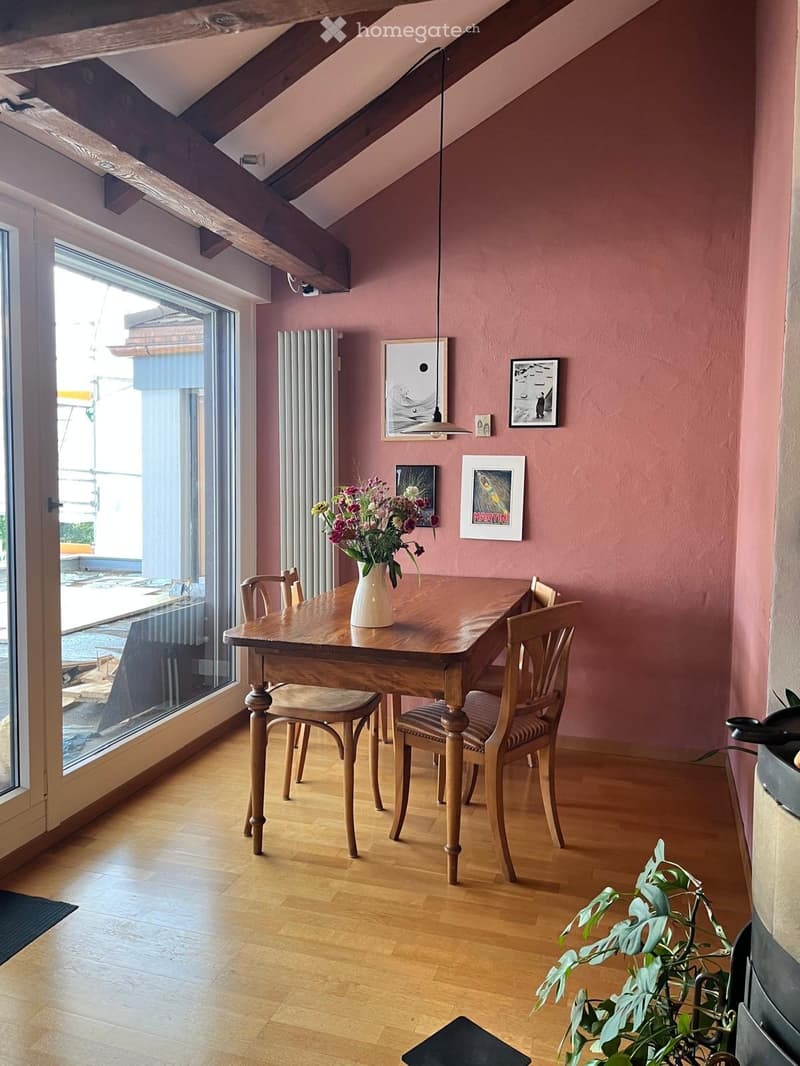 Dining area with wooden table, chairs, vase with flowers, and artwork on walls. Large window with view.