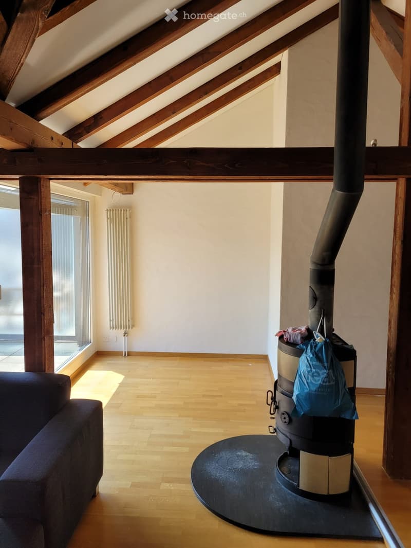Living room with wooden floor, exposed wooden beams, wood stove, sofa, radiator, and large window with blinds.