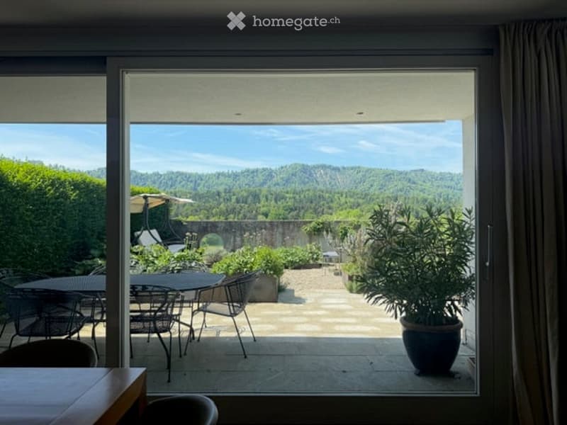 Balcony with large glass doors opening to a scenic outdoor area, featuring a table and chairs, potted plants, and a view of a landscaped garden and distant hills.