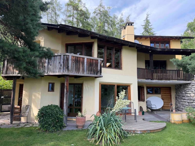 Two-story house with a wooden balcony, stone chimney, and outdoor seating area with a table and chairs.