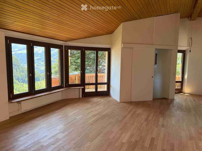 Empty room with wooden flooring, large windows, and a view of a forested area. The room has a slanted wooden ceiling and a bench near the windows.
