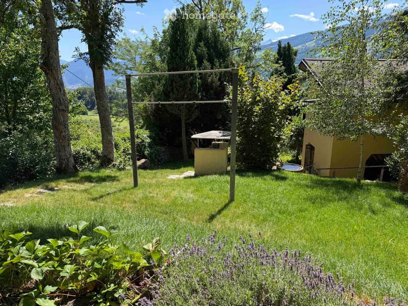 A well-maintained garden with green grass, lavender plants, trees, and a small yellow shed. The background features a scenic mountain view.