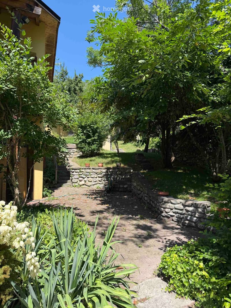 Garden path lined with stone walls, surrounded by lush greenery and trees, with a small building visible on the left.