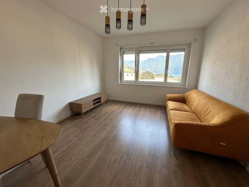 Modern living room with wooden flooring, large window offering a scenic mountain view, a beige sofa, a wooden table, and a small cabinet.