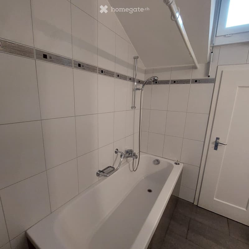 Modern bathroom with a bathtub, tiled walls, and a shower handle. Features include a handrail for support and a window for natural light.