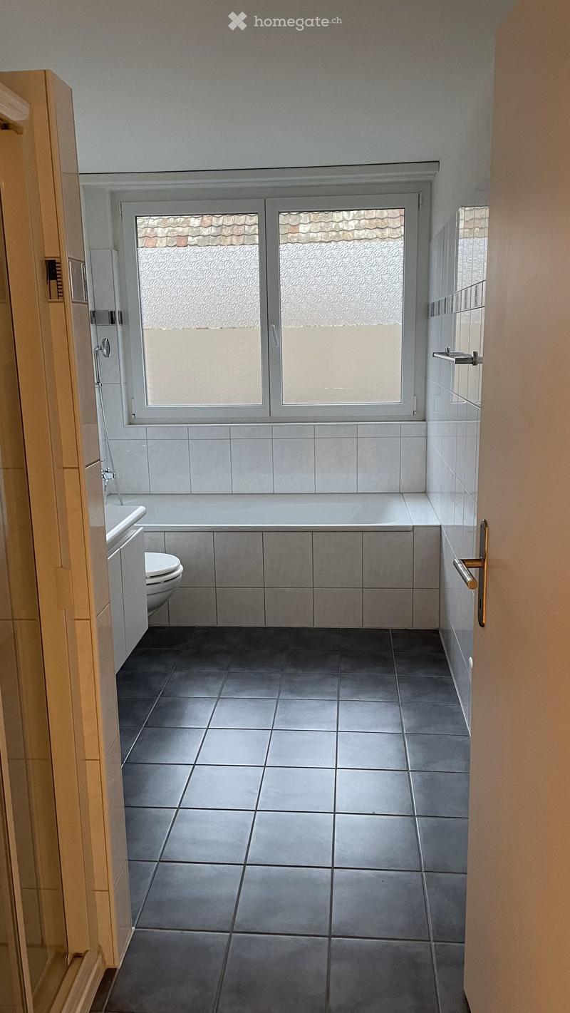 Modern bathroom with grey tiles, a bathtub, toilet, and a large window with frosted glass panels.