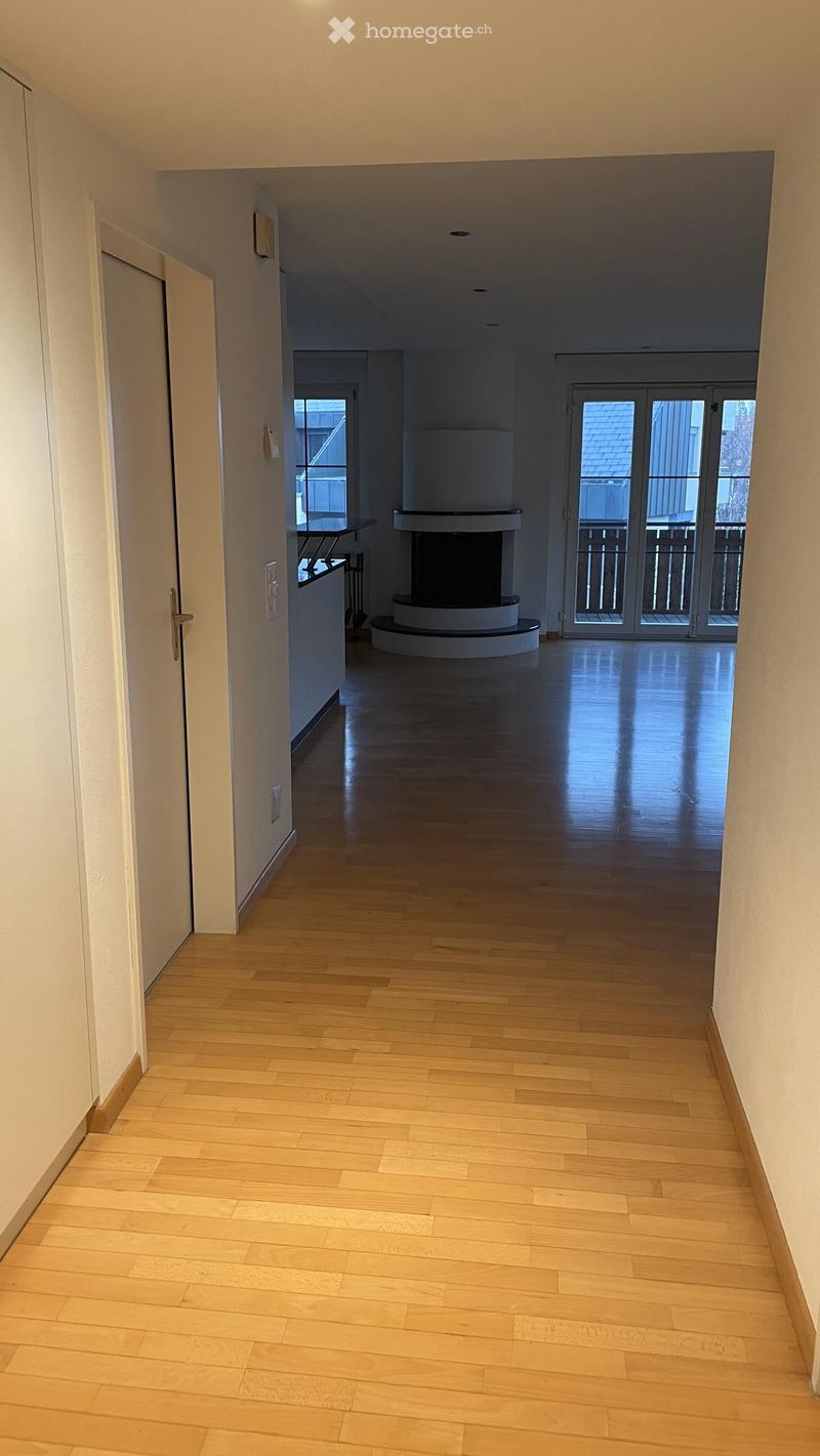 Empty hallway with light wooden flooring, white walls, and a central fireplace. A door on the left leads to another room, and a large window at the end provides natural light.