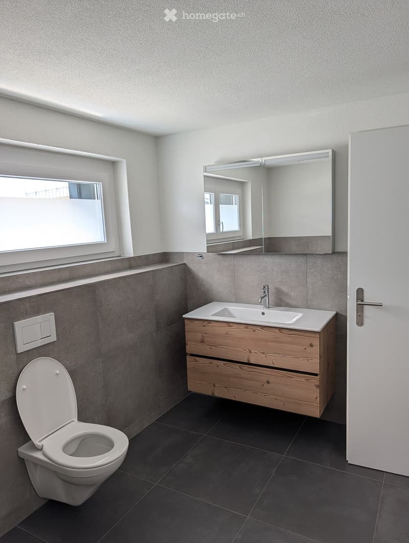 Modern bathroom with a white toilet, wooden sink cabinet, mirror, and dark tiled floor.
