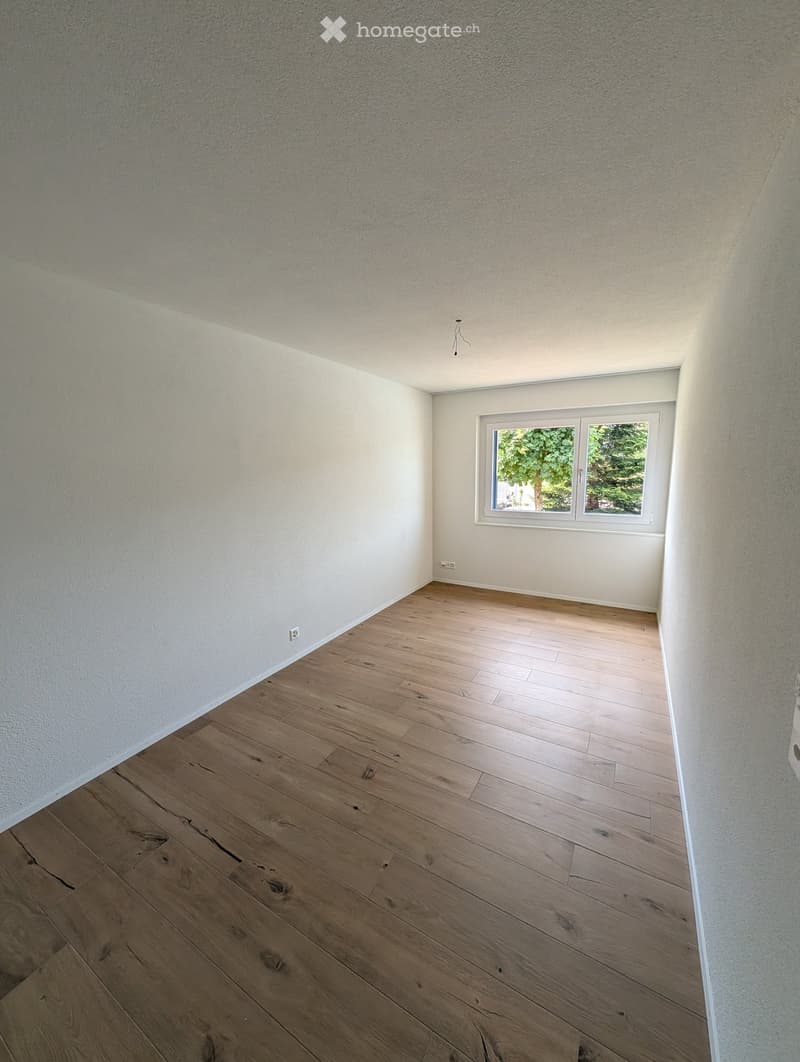Empty room with wooden flooring, white walls, and a window showing greenery outside.