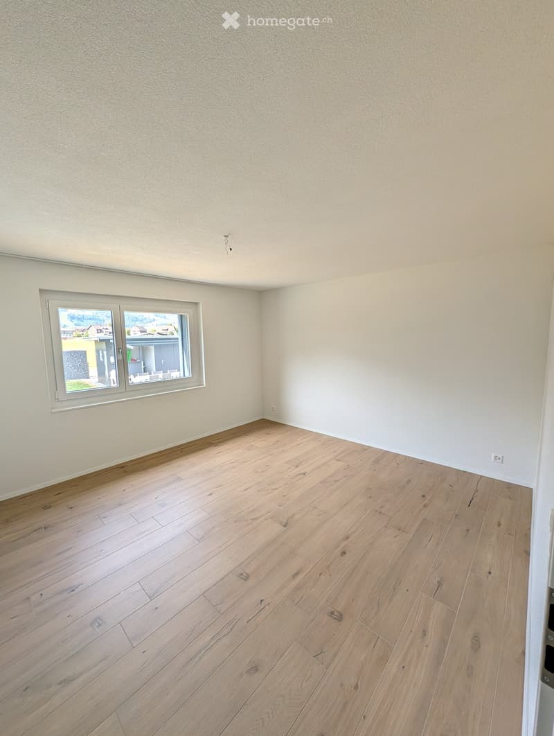 Empty room with light wooden flooring and white walls, featuring a large window with a view of the outside area.