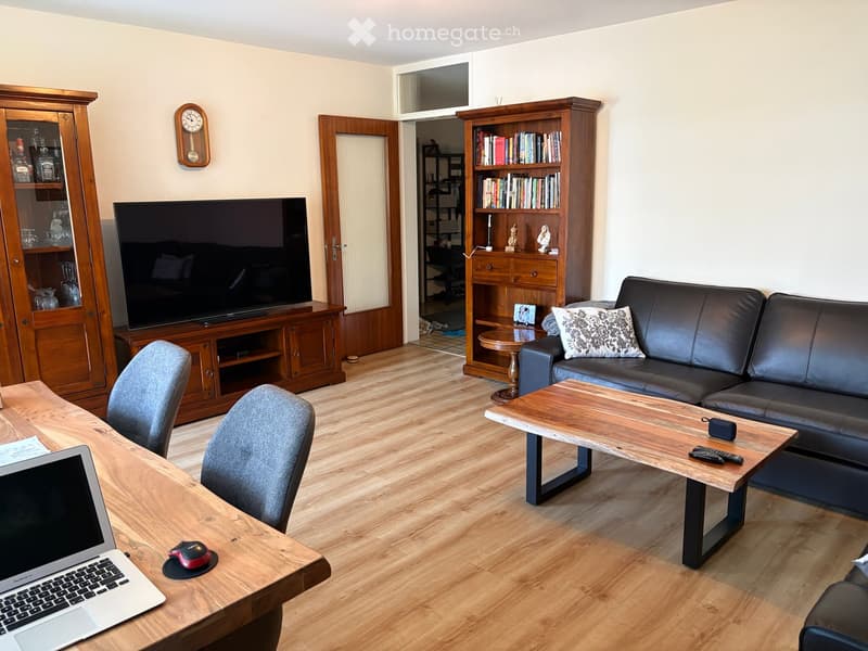 Living room with a wooden floor, a black leather sofa, a wooden coffee table, a laptop on a desk, and a TV on a wooden stand.